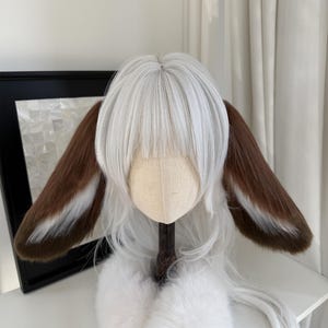 May include: A white wig featuring straight bangs and long layers, adorned with two large, plush animal ears. The ears are primarily brown with white fur accents along the inner edges, attached to the sides of the white synthetic hair fibers.