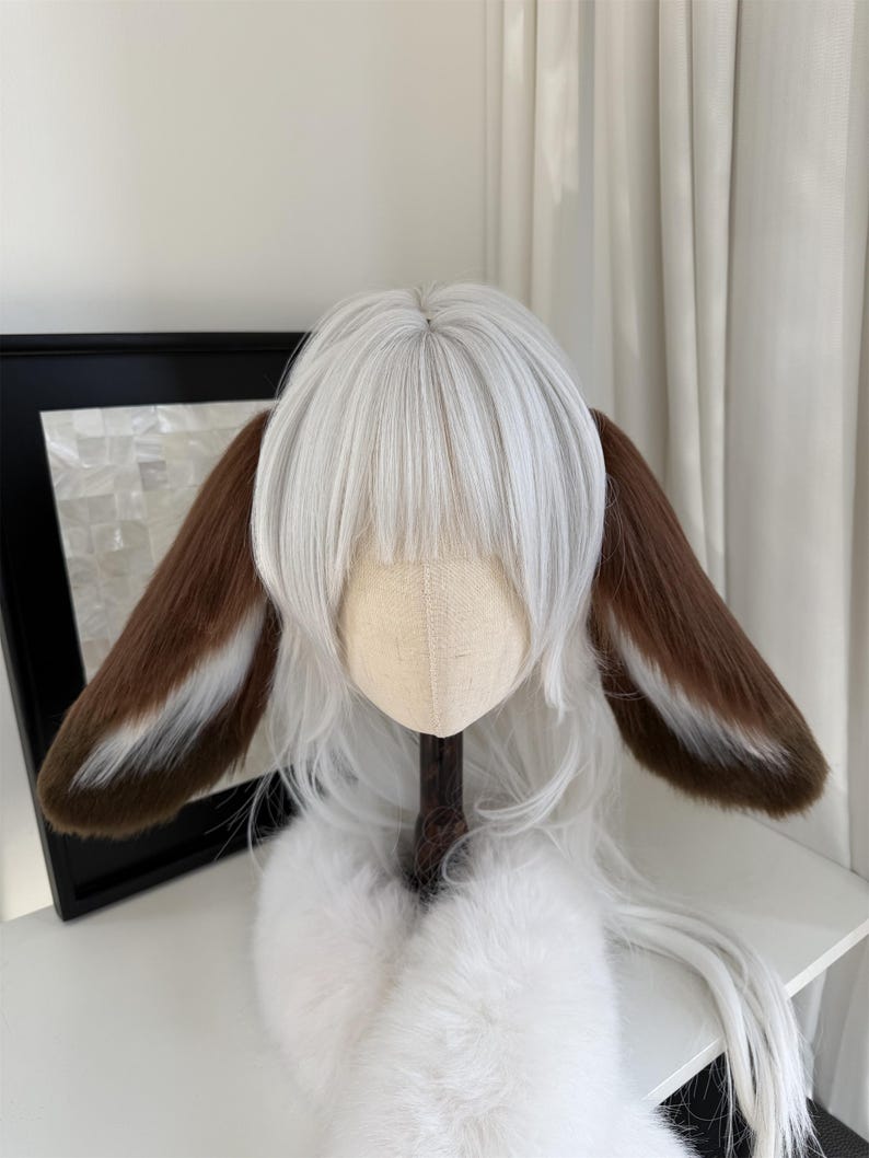 May include: A white wig featuring straight bangs and long layers, adorned with two large, plush animal ears. The ears are primarily brown with white fur accents along the inner edges, attached to the sides of the white synthetic hair fibers.