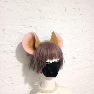 May include: A headband featuring large, rounded animal ears with light pink inner sections and brown fur outer sections. The ears are positioned on a mannequin head with short, layered, light brown hair, a black face mask, and a white beaded necklace.