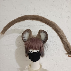 May include: A set of faux fur animal ears and a long, curved faux fur tail. The ears are brown with white inner fur and are attached to a headband. The tail is long, tapered, and brown, matching the color of the ears. The set is displayed on a mannequin.
