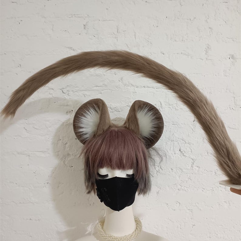 May include: A set of faux fur animal ears and a long, curved faux fur tail. The ears are brown with white inner fur and are attached to a headband. The tail is long, tapered, and brown, matching the color of the ears. The set is displayed on a mannequin.