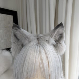 Puede incluir: Un par de orejas de lobo de piel sint&eacute;tica gris y blanca unidas a una peluca. Las orejas est&aacute;n erguidas con puntas grises m&aacute;s oscuras y esponjosas, mientras que la peluca tiene cabello liso de color gris claro.