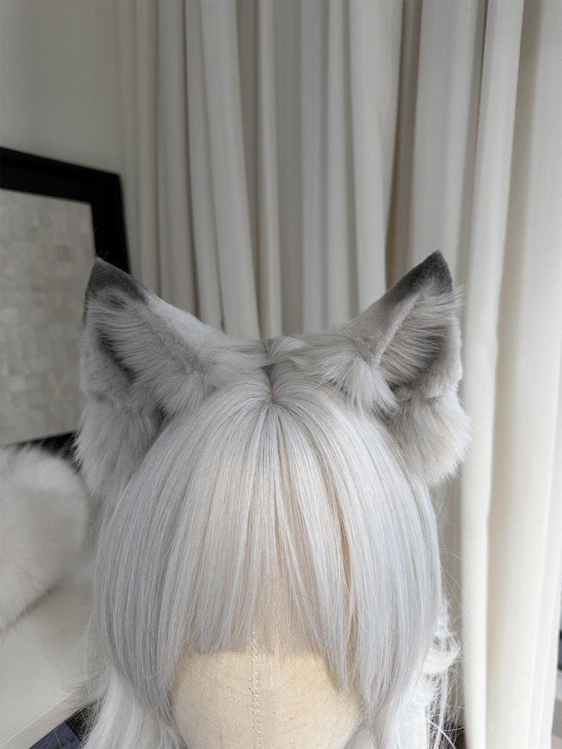 Puede incluir: Un par de orejas de lobo de piel sint&eacute;tica gris y blanca unidas a una peluca. Las orejas est&aacute;n erguidas con puntas grises m&aacute;s oscuras y esponjosas, mientras que la peluca tiene cabello liso de color gris claro.