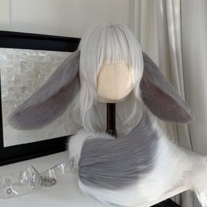 May include: A white wig featuring straight hair with bangs and two large, long, grey and white faux fur animal ears attached to the sides. The wig is displayed on a mannequin head, accompanied by a matching grey and white faux fur tail.