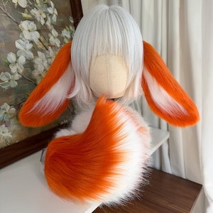 May include: A white wig with straight bangs displayed on a mannequin head, featuring two long, plush, orange and white animal ears attached to the sides and a large, fluffy, orange and white faux fur tail positioned in front of the base.