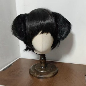 May include: A short, straight black wig featuring integrated plush animal ears on both sides. The wig is styled with straight bangs and is displayed on a neutral-colored mannequin head mounted on a dark wooden stand with a circular base.