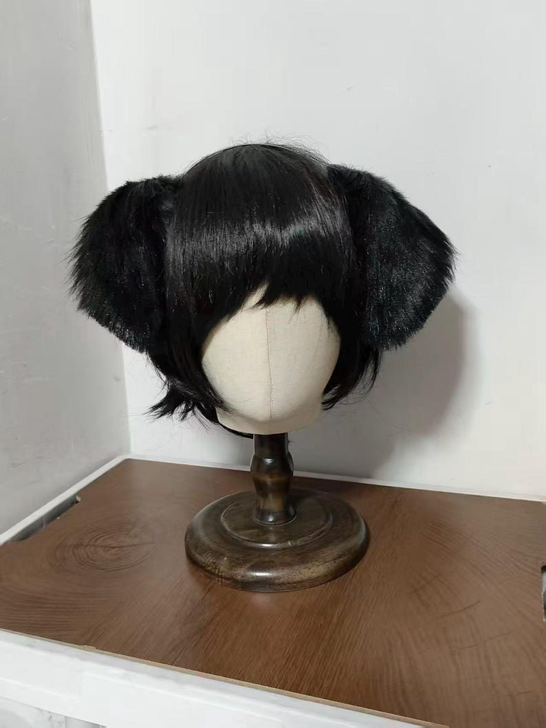May include: A short, straight black wig featuring integrated plush animal ears on both sides. The wig is styled with straight bangs and is displayed on a neutral-colored mannequin head mounted on a dark wooden stand with a circular base.
