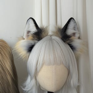 May include: A pair of faux fur fox ears with black tips, white inner ears, and a mix of tan and gray fur. The ears are attached to a headband, displayed on a mannequin head with white hair.