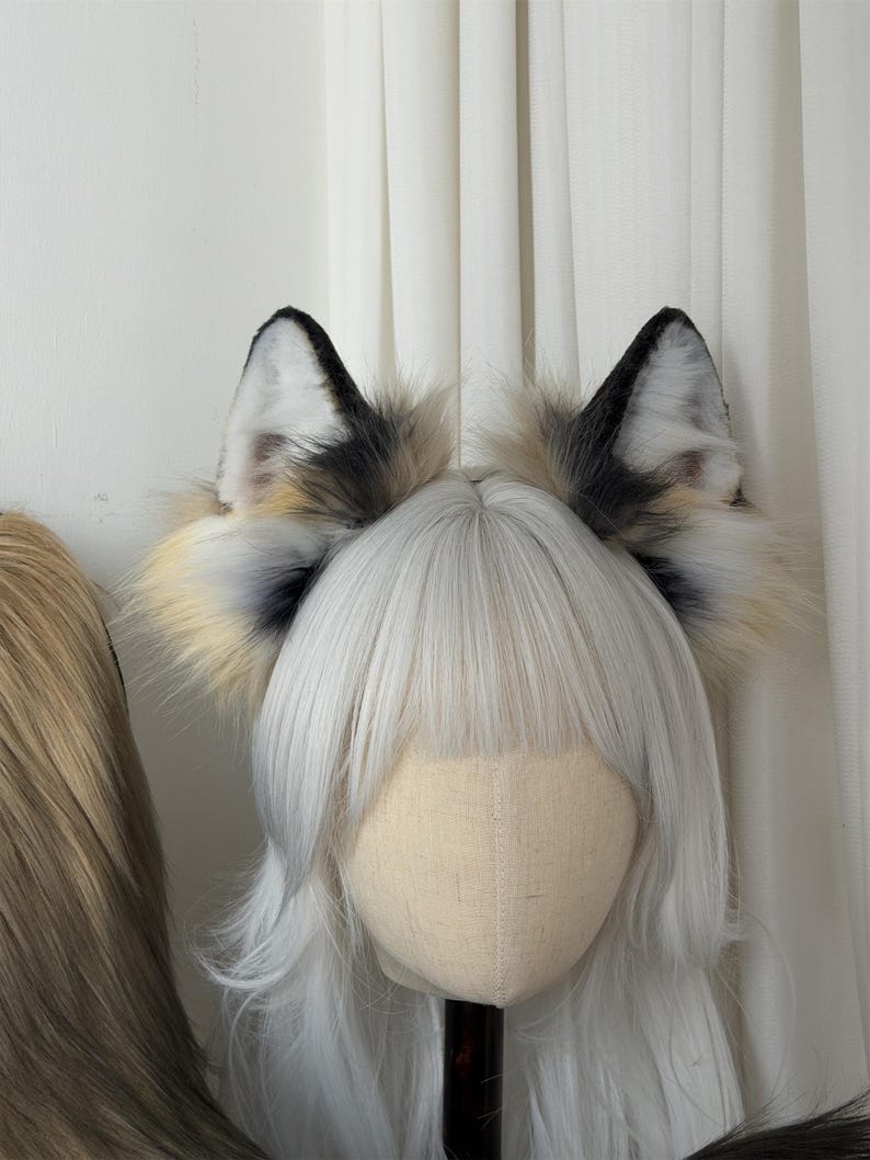 May include: A pair of faux fur fox ears with black tips, white inner ears, and a mix of tan and gray fur. The ears are attached to a headband, displayed on a mannequin head with white hair.