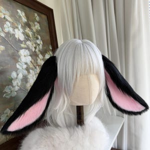 May include: A white wig with straight bangs and long layers, featuring two large, plush animal ears attached to the sides. The ears have black outer fur and soft pink inner fur. A white faux fur collar is positioned around the base of the wig.