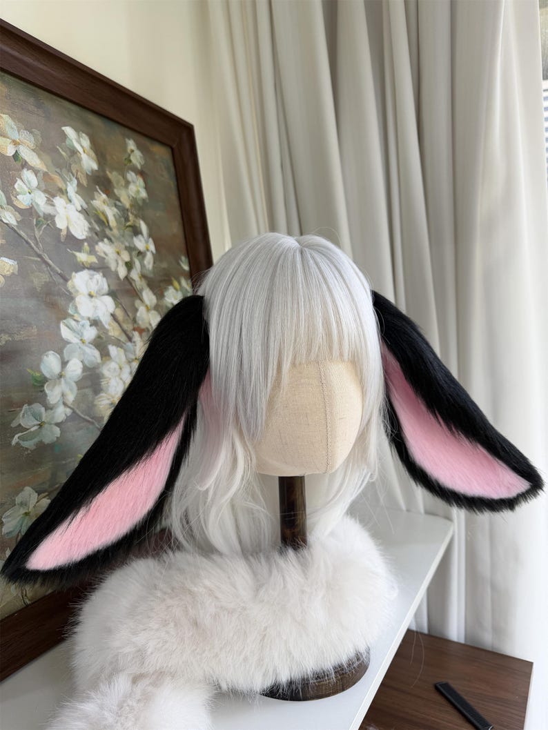 May include: A white wig with straight bangs and long layers, featuring two large, plush animal ears attached to the sides. The ears have black outer fur and soft pink inner fur. A white faux fur collar is positioned around the base of the wig.