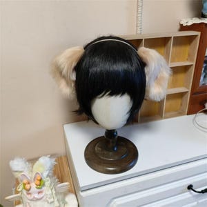 May include: A black wig styled with short, straight hair and a thin headband featuring two large, fluffy, light beige faux fur animal ears attached to the sides. The wig is displayed on a neutral-colored mannequin head with a dark wooden base.