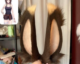 May include: A pair of tall, pointed animal ears attached to a thin metal headband. The ears are covered in dark brown faux fur with lighter tan fur on the inner surfaces and tips, mimicking the appearance of a rabbit or hare-like creature.