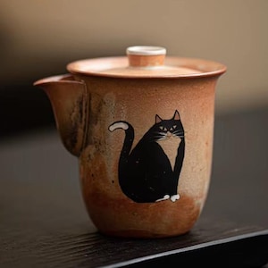 Op de afbeelding: Een bruine theepot van keramiek met een zwart kattenontwerp. De kat heeft witte poten en een witte vlek op zijn borst. De theepot heeft een deksel en een tuit.