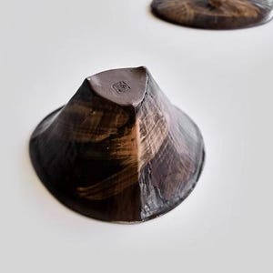 Pu&ograve; includere: Una ciotola in ceramica marrone scuro con un design testurizzato che ricorda le pennellate. La ciotola ha una forma angolare unica e un piccolo segno quadrato all'interno.