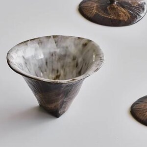 Pu&ograve; includere: Una tazza da t&egrave; in ceramica bianca e marrone con un bordo largo e svasato e una base conica. La tazza ha una smaltatura macchiata con un interno bianco.