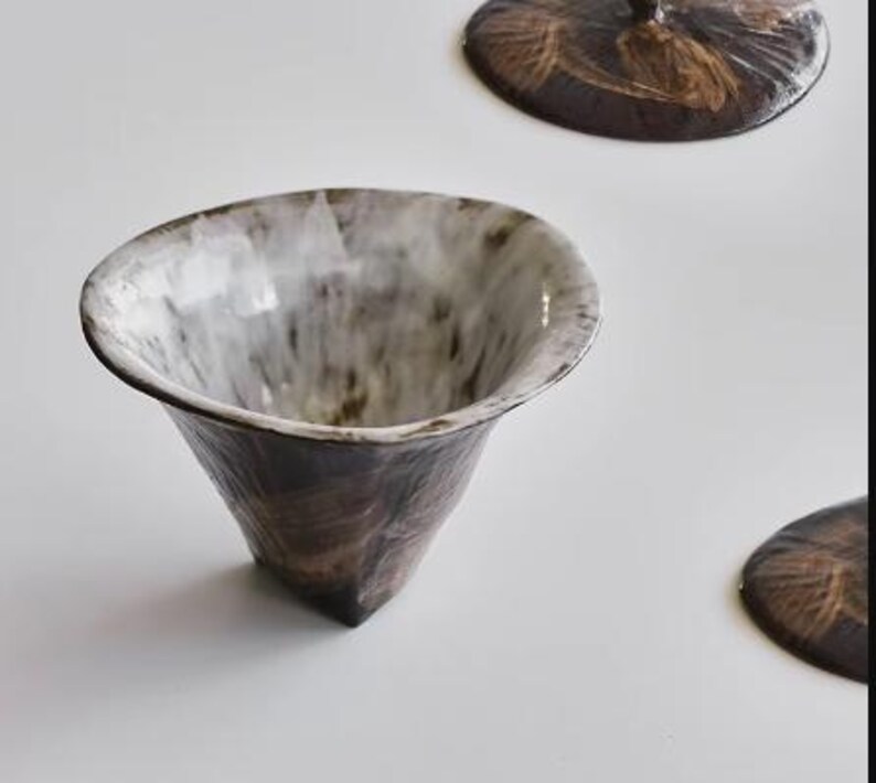 Pu&ograve; includere: Una tazza da t&egrave; in ceramica bianca e marrone con un bordo largo e svasato e una base conica. La tazza ha una smaltatura macchiata con un interno bianco.