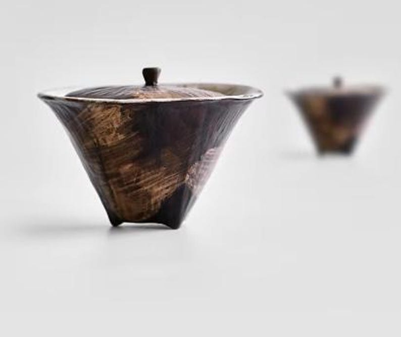 Pu&ograve; includere: Una tazza da t&egrave; in ceramica marrone con un coperchio e tre gambe. La tazza da t&egrave; ha un design testurizzato, simile a pennellate.