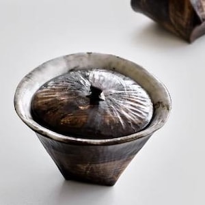 Pu&ograve; includere: Una ciotola in ceramica con un coperchio. La ciotola &egrave; bianca con una glassa nera e marrone. Il coperchio ha una superficie testurizzata e un piccolo pomo in cima.