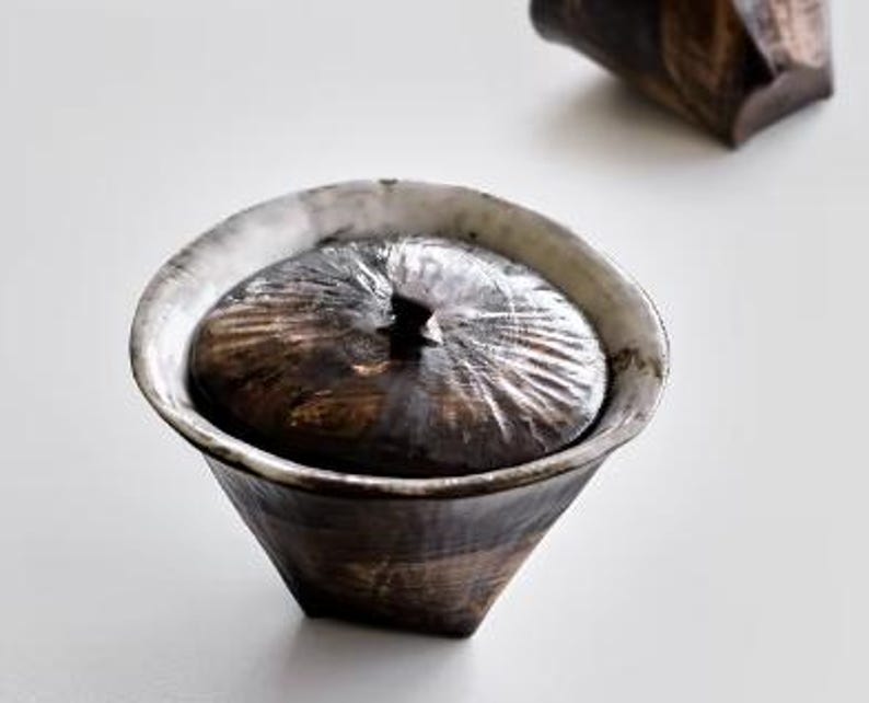 Pu&ograve; includere: Una ciotola in ceramica con un coperchio. La ciotola &egrave; bianca con una glassa nera e marrone. Il coperchio ha una superficie testurizzata e un piccolo pomo in cima.