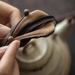 Puede incluir: Una funda para tetera de tela marrón con una banda elástica negra y un botón marrón. La funda se coloca sobre una tetera de cerámica.