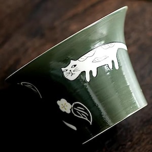 Könnte beinhalten: Eine grüne Keramikschüssel mit einem weißen Katzen- und Blumendesign. Die Schüssel ist glasiert und hat eine glänzende Oberfläche.