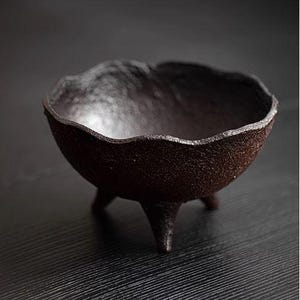 Puede incluir: Un cuenco de cerámica marrón oscuro con una superficie texturizada y tres patas. El cuenco está ligeramente acampanado en la parte superior y tiene un fondo redondeado.