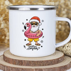 Puede incluir: Taza de esmalte blanco con una ilustraci&oacute;n de dibujos animados de Pap&aacute; Noel con gafas de sol y un gorro rojo, sujetando un flotador de flamenco rosa. El texto "merry christmas" est&aacute; escrito debajo de la imagen.