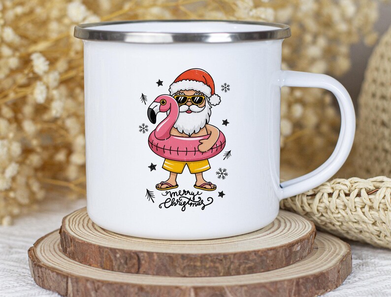 Puede incluir: Taza de esmalte blanco con una ilustraci&oacute;n de dibujos animados de Pap&aacute; Noel con gafas de sol y un gorro rojo, sujetando un flotador de flamenco rosa. El texto "merry christmas" est&aacute; escrito debajo de la imagen.