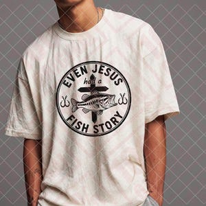 May include: Cream-colored t-shirt featuring a circular graphic with the text "EVEN JESUS had a FISH STORY." The graphic includes a fish, cross, and fishing hooks. The shirt is a casual, short-sleeved design.