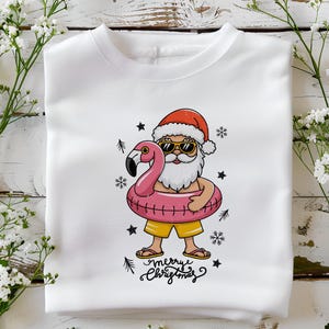 Puede incluir: Sudadera blanca con una ilustraci&oacute;n de dibujos animados de Pap&aacute; Noel con gafas de sol y un gorro de Pap&aacute; Noel, montando un flotador de flamenco rosa. El texto "Merry Christmas" est&aacute; escrito debajo de la imagen.