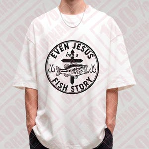 May include: White t-shirt with a black graphic. The graphic features a fish, cross, and the text "EVEN JESUS had a FISH STORY" in a circular design. The shirt has short sleeves and a relaxed fit.
