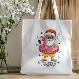 Puede incluir: Bolsa de tela blanca con una ilustraci&oacute;n de dibujos animados de Pap&aacute; Noel con gafas de sol montando un flotador de flamenco rosa. El texto "Merry Christmas" est&aacute; impreso debajo de la imagen.