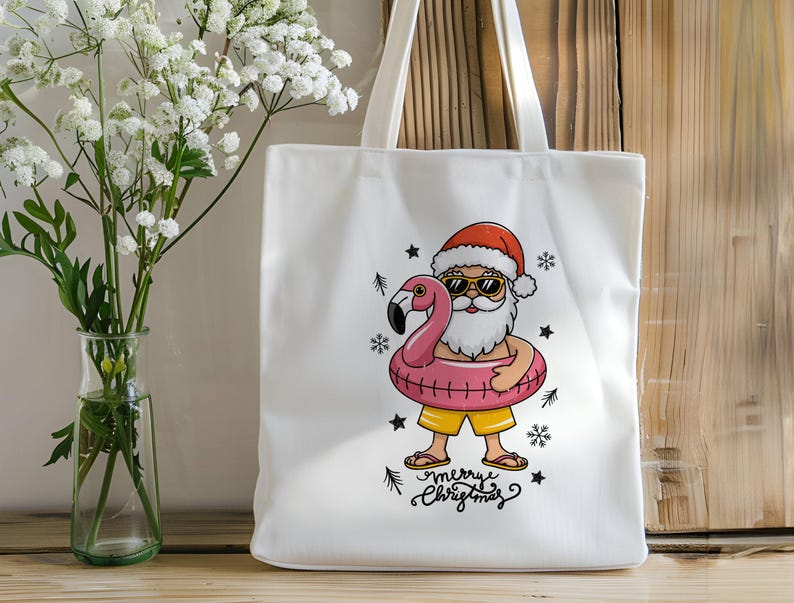 Puede incluir: Bolsa de tela blanca con una ilustraci&oacute;n de dibujos animados de Pap&aacute; Noel con gafas de sol montando un flotador de flamenco rosa. El texto "Merry Christmas" est&aacute; impreso debajo de la imagen.