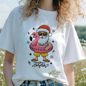 Puede incluir: Camiseta blanca con un gr&aacute;fico de Santa Claus con gafas de sol montando un flotador de flamenco rosa. El texto "Merry Christmas" est&aacute; impreso debajo del gr&aacute;fico.