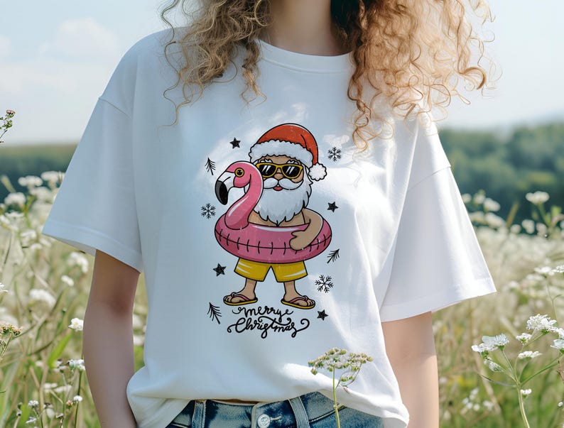 Puede incluir: Camiseta blanca con un gr&aacute;fico de Santa Claus con gafas de sol montando un flotador de flamenco rosa. El texto "Merry Christmas" est&aacute; impreso debajo del gr&aacute;fico.