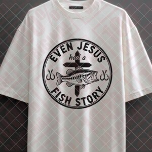 May include: White t-shirt with a black graphic. The graphic is a circular design with the words "EVEN JESUS had a FISH STORY" around a cross and a fish. The shirt has short sleeves and a crew neck.