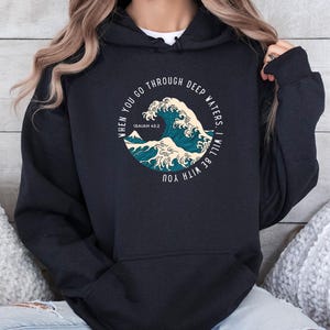 K&ouml;nnte beinhalten: Schwarzer Hoodie mit einem Grafikdruck einer Welle und dem Text "When you go through deep waters, I will be with you. Isaiah 43:2"