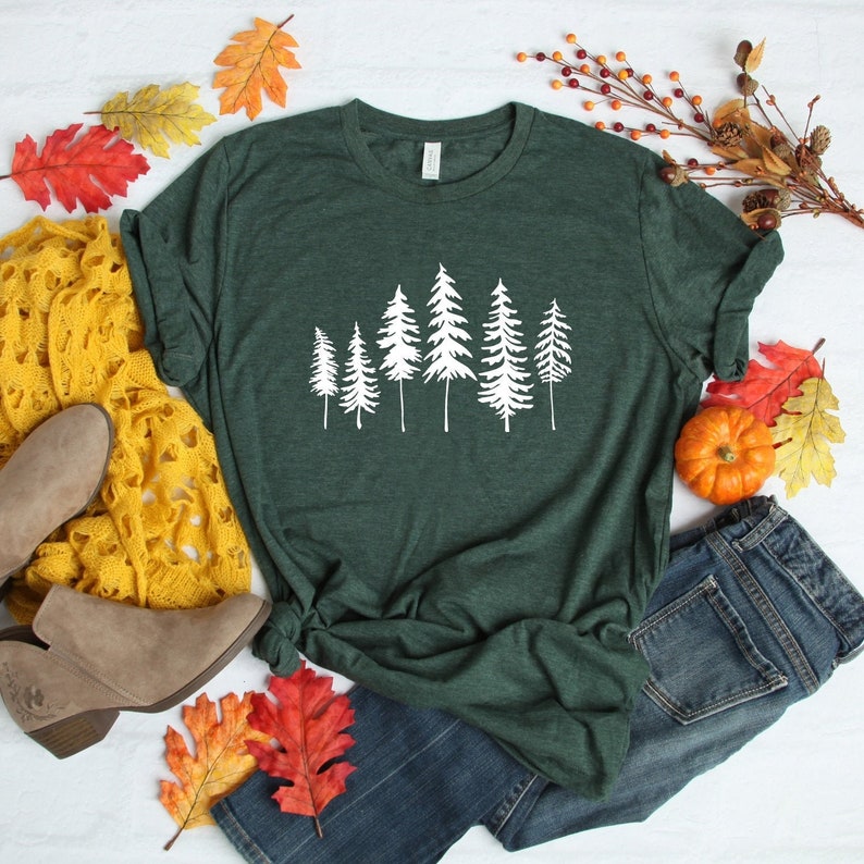 May include: A dark green t-shirt featuring a white graphic of a row of stylised pine trees. The shirt is laid out on a white surface with a pair of brown ankle boots, a yellow crocheted scarf, blue jeans, and autumn leaves.