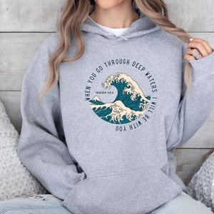 K&ouml;nnte beinhalten: Grauer Kapuzenpullover mit einem Grafikdruck einer blauen Welle und dem Text "When you go through deep waters, I will be with you. Isaiah 43:2"