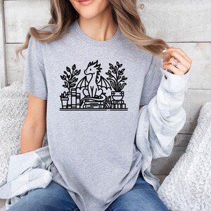 May include: A gray t-shirt with a black and white illustration of a dragon sitting on a stack of books with plants in pots on either side.