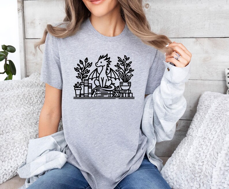May include: A gray t-shirt with a black and white illustration of a dragon sitting on a stack of books with plants in pots on either side.