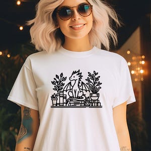 May include: White t-shirt with a black line drawing of a dragon sitting on a stack of books with plants around it.