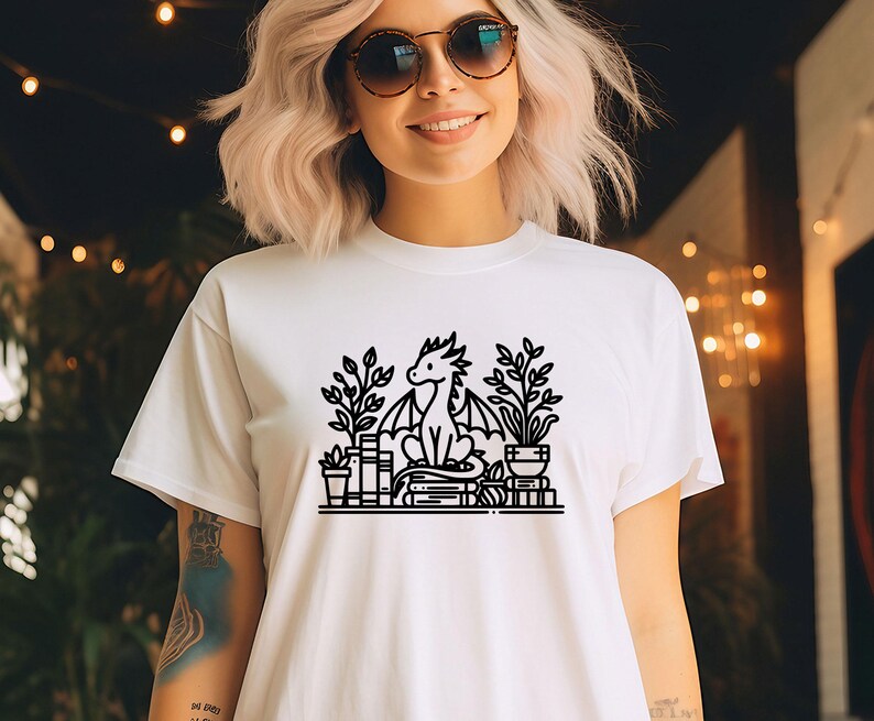 May include: White t-shirt with a black line drawing of a dragon sitting on a stack of books with plants around it.
