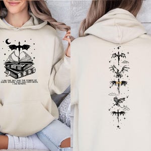 May include: A cream-coloured hooded sweatshirt. The front displays a graphic of books, a dragon, and the text "I AM THE SKY AND THE POWER OF EVERY STORM THAT HAS EVER BEEN. I AM INFINITE." The back has dragon illustrations.
