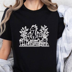 May include: Black t-shirt with a white graphic of a dragon sitting on a stack of books and surrounded by plants.
