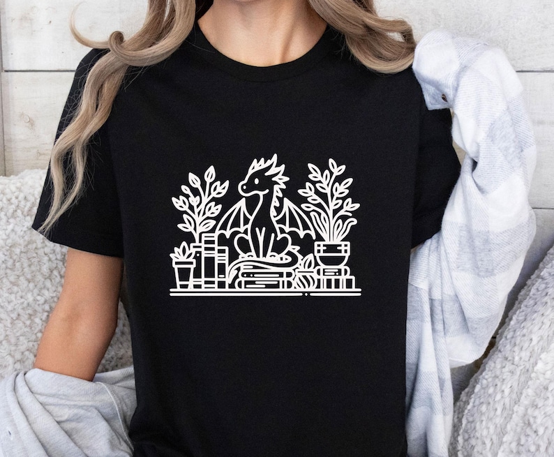 May include: Black t-shirt with a white graphic of a dragon sitting on a stack of books and surrounded by plants.