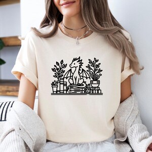 May include: A white t-shirt with a black line drawing of a dragon sitting on a stack of books with plants in pots on either side. The dragon has a curled tail and is looking to the left.
