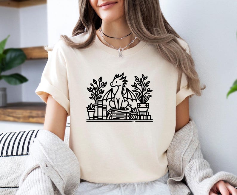 May include: A white t-shirt with a black line drawing of a dragon sitting on a stack of books with plants in pots on either side. The dragon has a curled tail and is looking to the left.