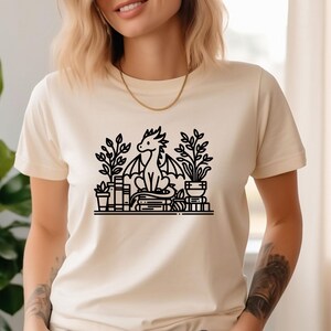 May include: A white t-shirt with a black line drawing of a dragon sitting on a stack of books with plants around it.
